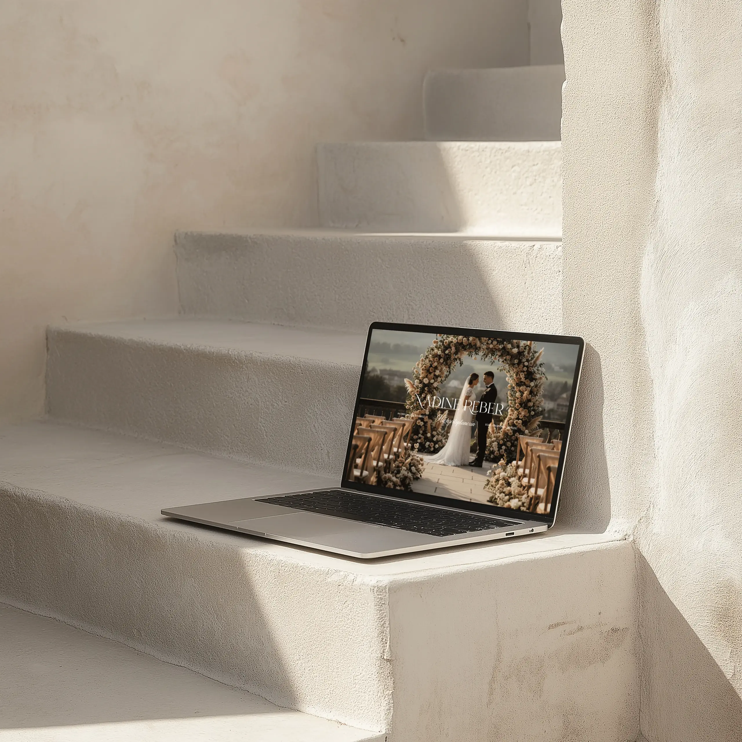 Laptop auf hellen Stufen zeigt eine Website mit einem Hochzeitspaar unter einem Blumenbogen. Das Bild vermittelt Stil, Ruhe und Hochwertigkeit.